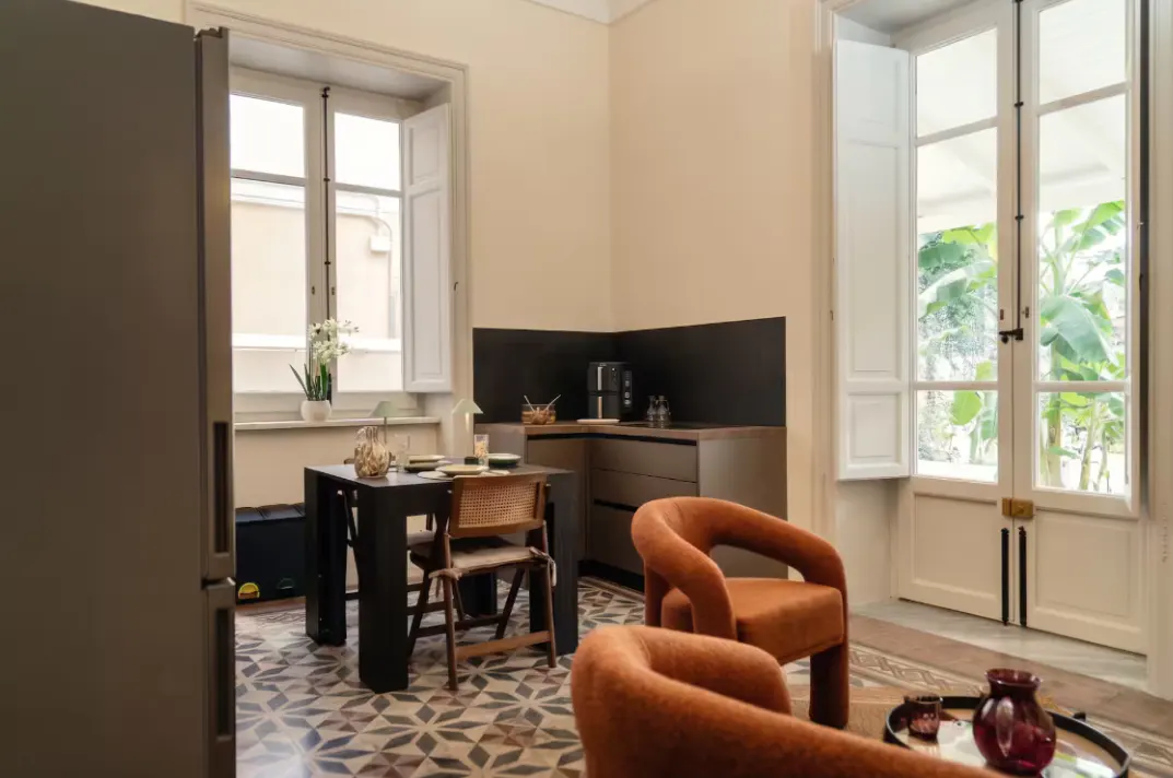 Interior view of a modern apartment with a black table, brown chairs, and orange armchairs. White windows with open shutters let in natural light.
