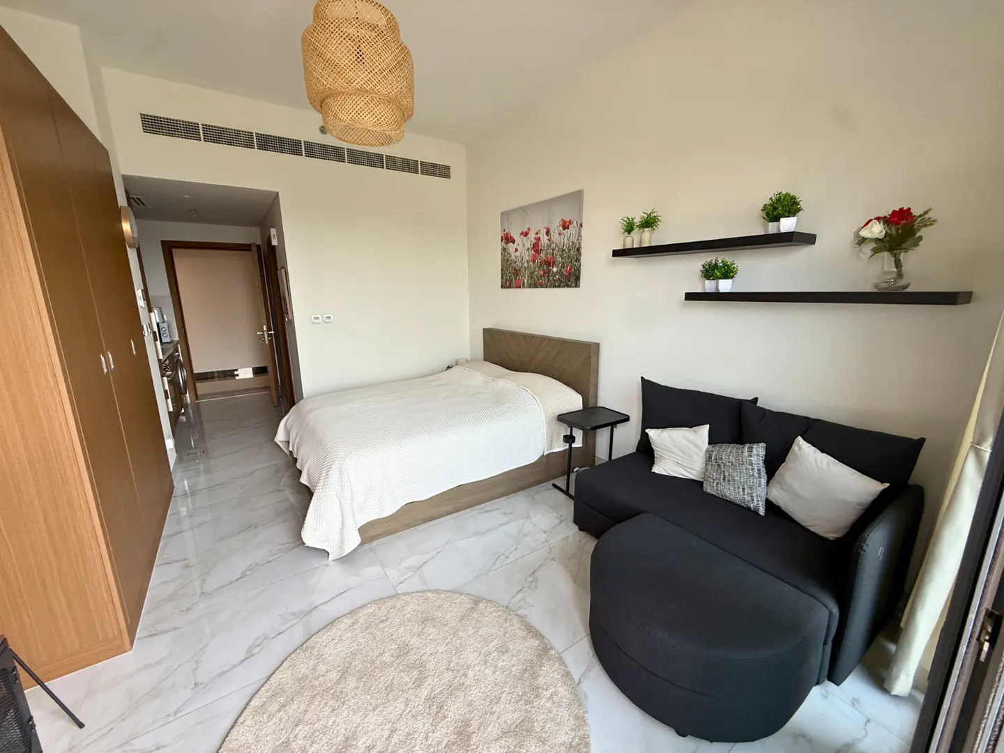 A studio apartment with a bed, black sofa, and light wood cabinets. Two shelves with plants are on the wall.