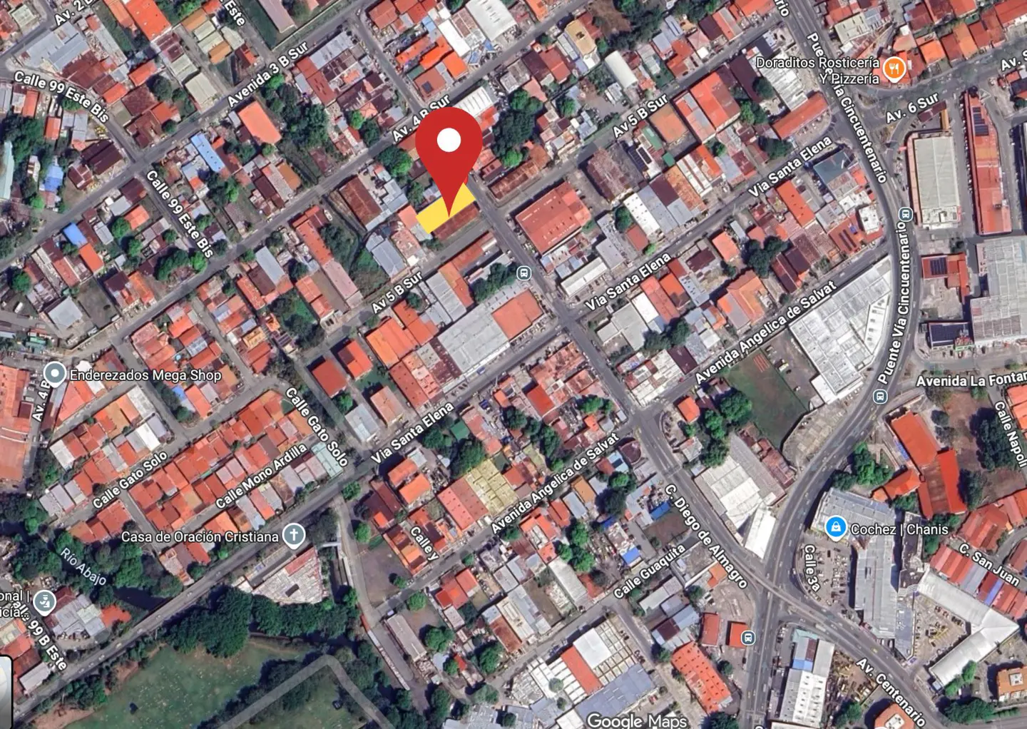 Aerial view of a neighborhood with a red map marker. Streets, buildings with red roofs, and some green trees are visible.