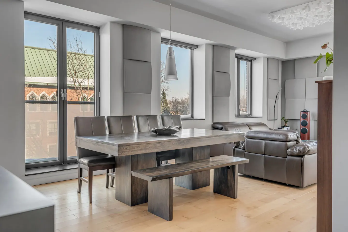 A modern dining room with a large wooden table, leather chairs, and a bench. Windows offer a view of a building and trees. A leather sofa is visible in the background.