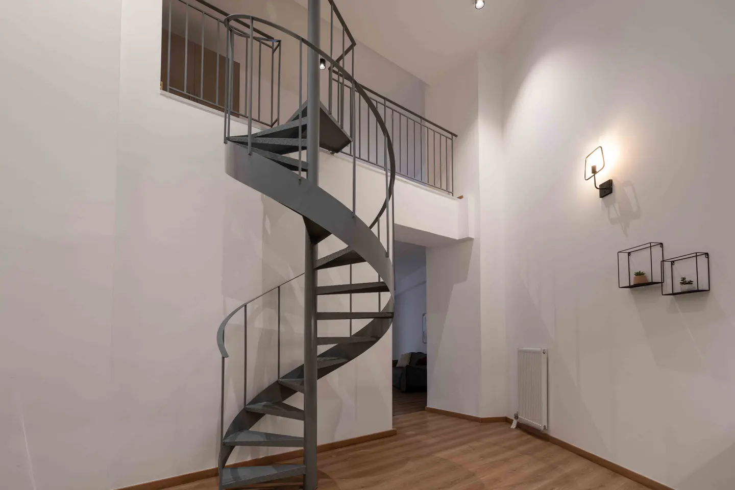 Interior view of a room with a gray spiral staircase, white walls, and light wood floors. A modern light fixture and shelves are on the wall.