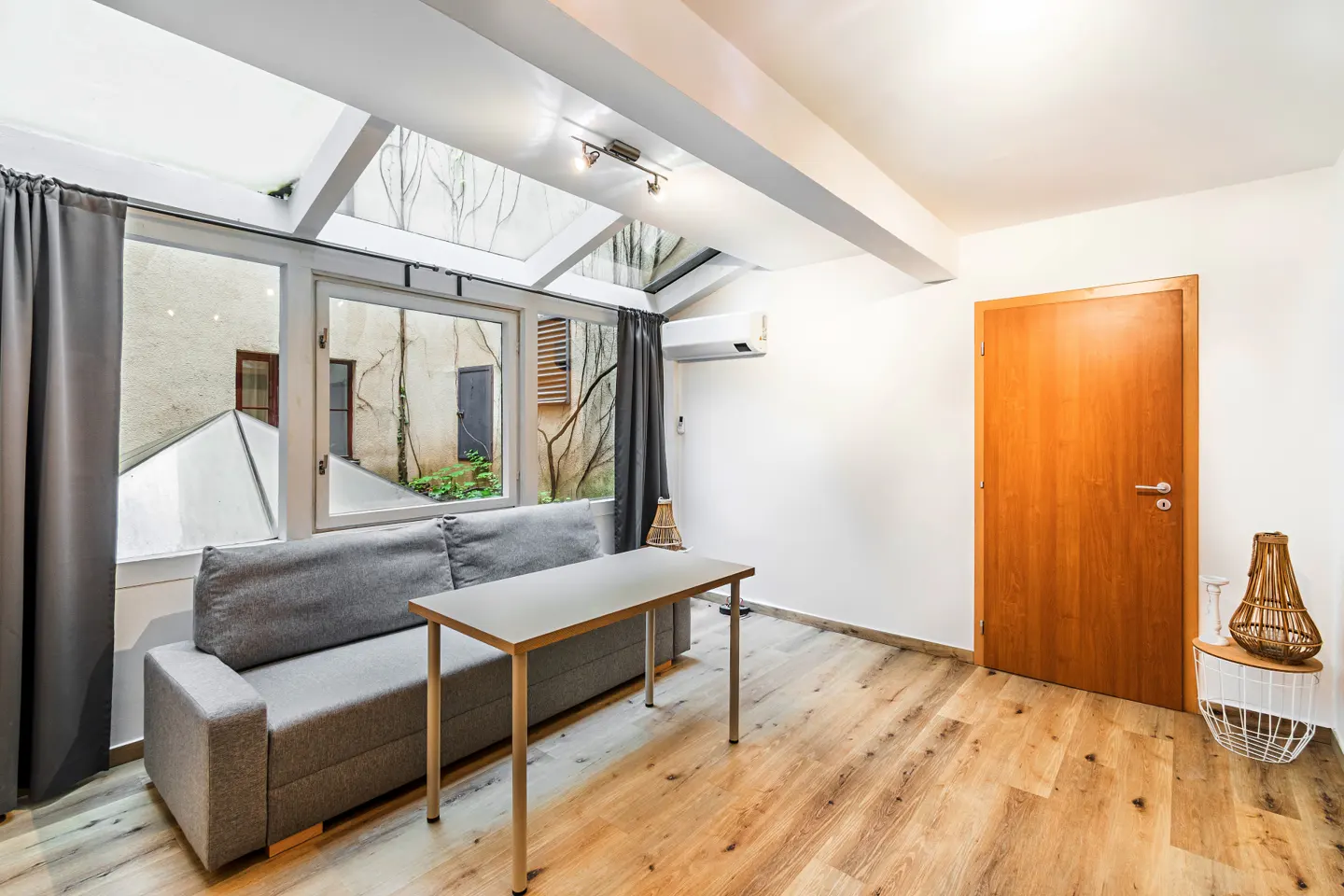 Modern room with a grey sofa, a simple table, wood floors, a skylight, and a wooden door leading to an outdoor courtyard.
