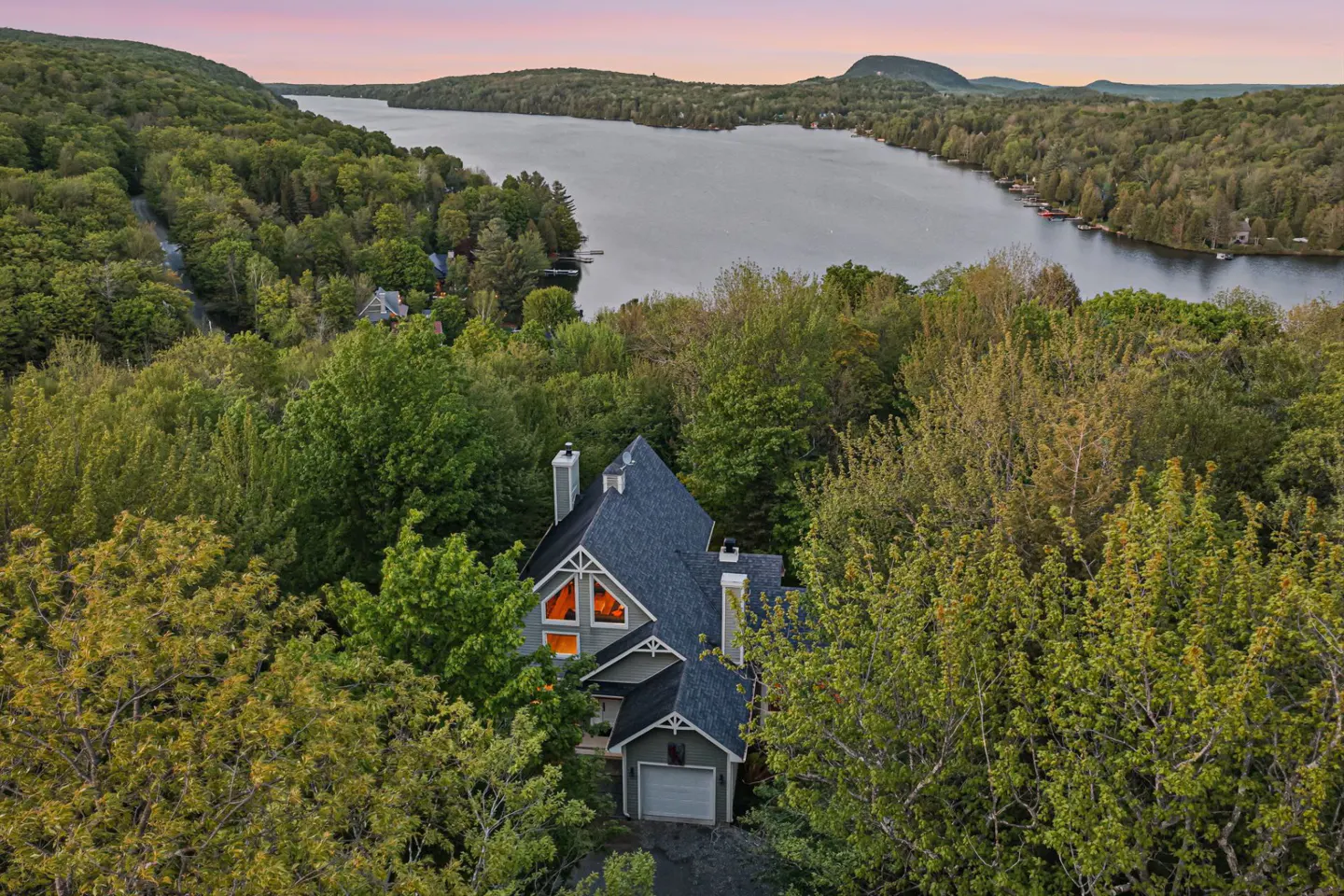 Aerial view of a house nestled in a lush forest overlooking a serene lake at sunset with a pink and orange sky.