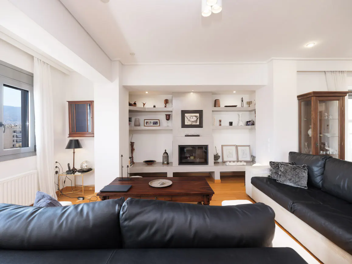 Bright living room with black leather sofas, a wooden coffee table, and a fireplace with built-in shelves. A window offers a city view.