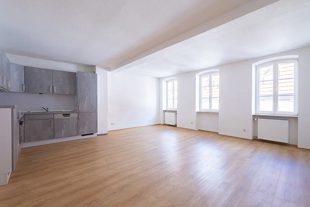 Bright, empty apartment with wood floors, white walls, and gray kitchen cabinets. Three windows let in natural light.