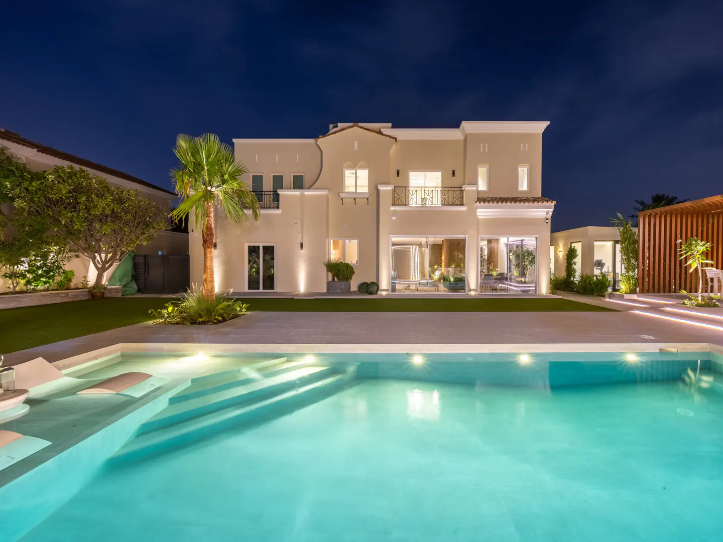 Night view of a two-story beige house with a pool. The pool has built-in lounge chairs and steps. A palm tree stands near the house.