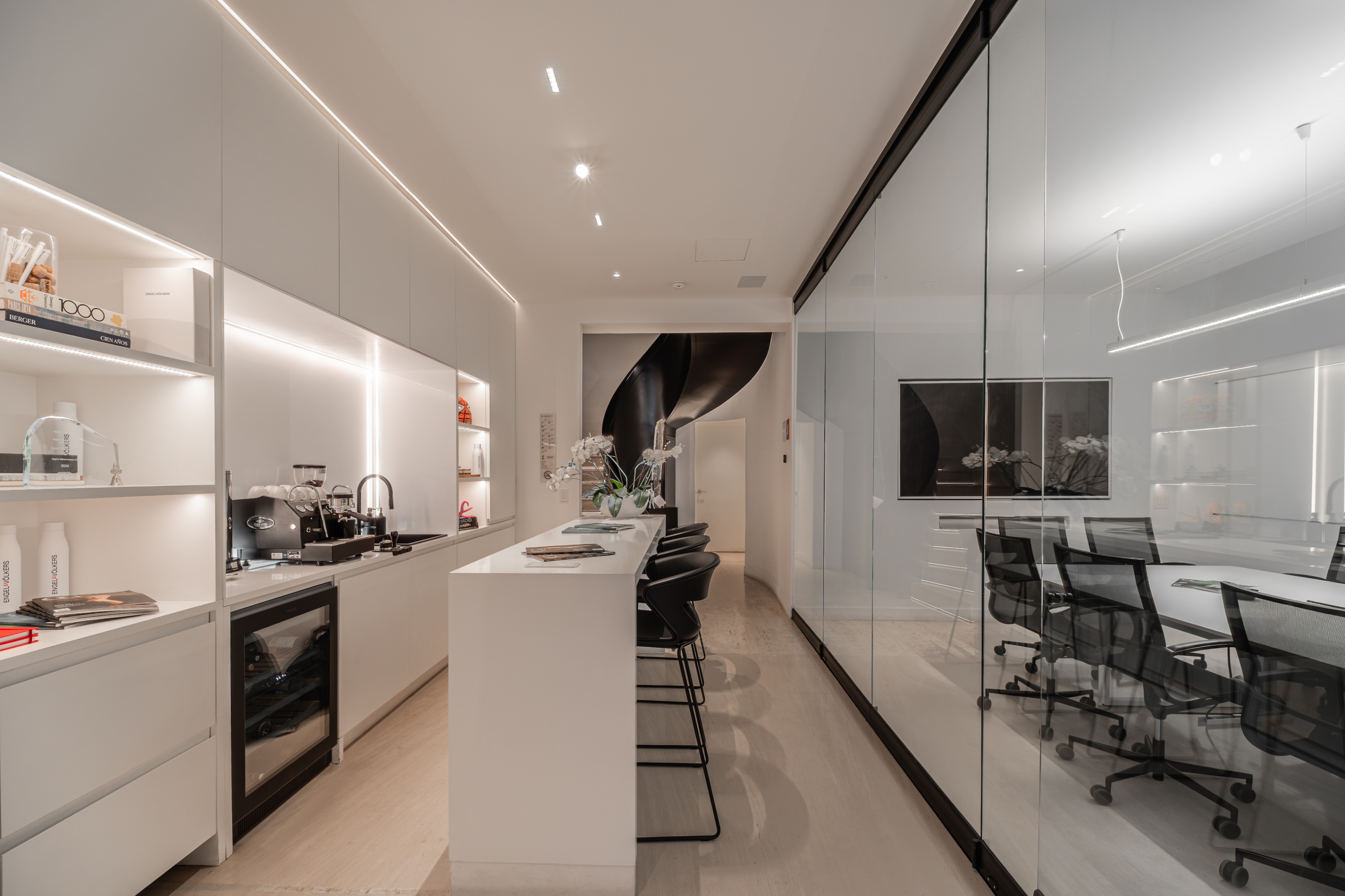 Modern office kitchen with white cabinets, a central island with bar stools, and a glass-walled conference room.