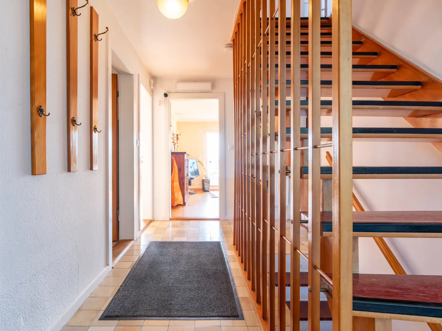 Hallway with wooden stairs, coat rack, and a gray rug on a tiled floor. A doorway leads to a bright room.