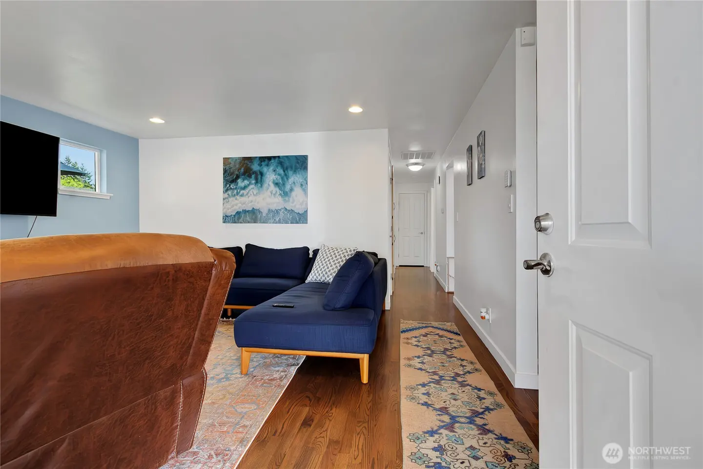 Living room with blue sectional, brown leather chair, and ocean art. Hallway with white door and patterned rug.
