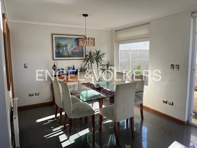 Bright dining room with a glass table, six chairs, and a patterned runner. A painting hangs above a sideboard with decorative items. A window provides natural light.