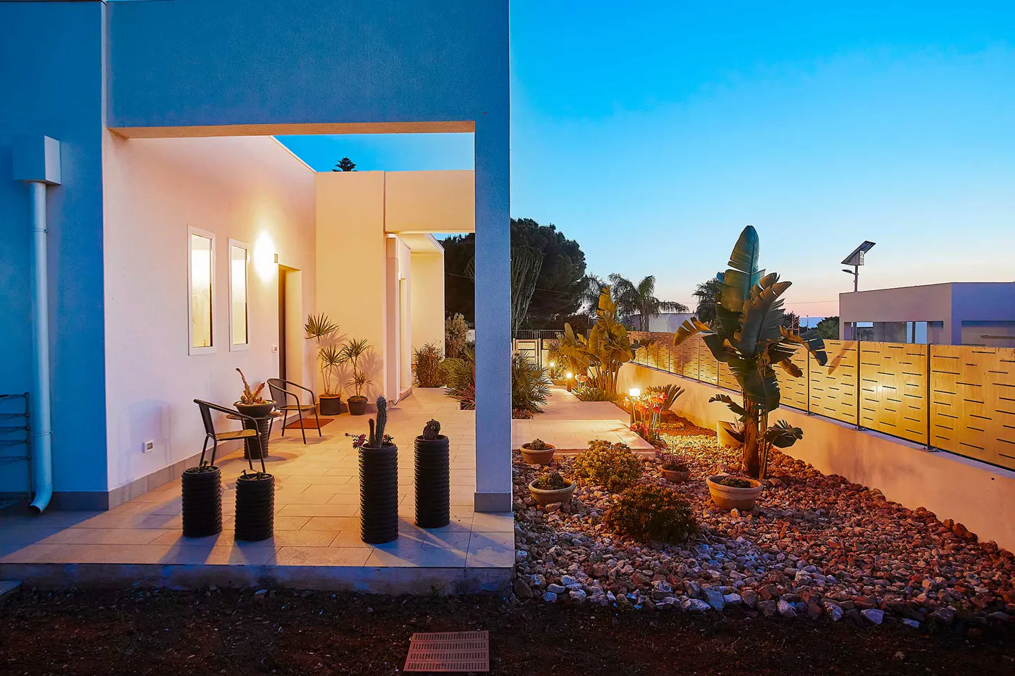 Exterior view of a modern home with a patio, chairs, and a garden with plants and a fence at dusk.