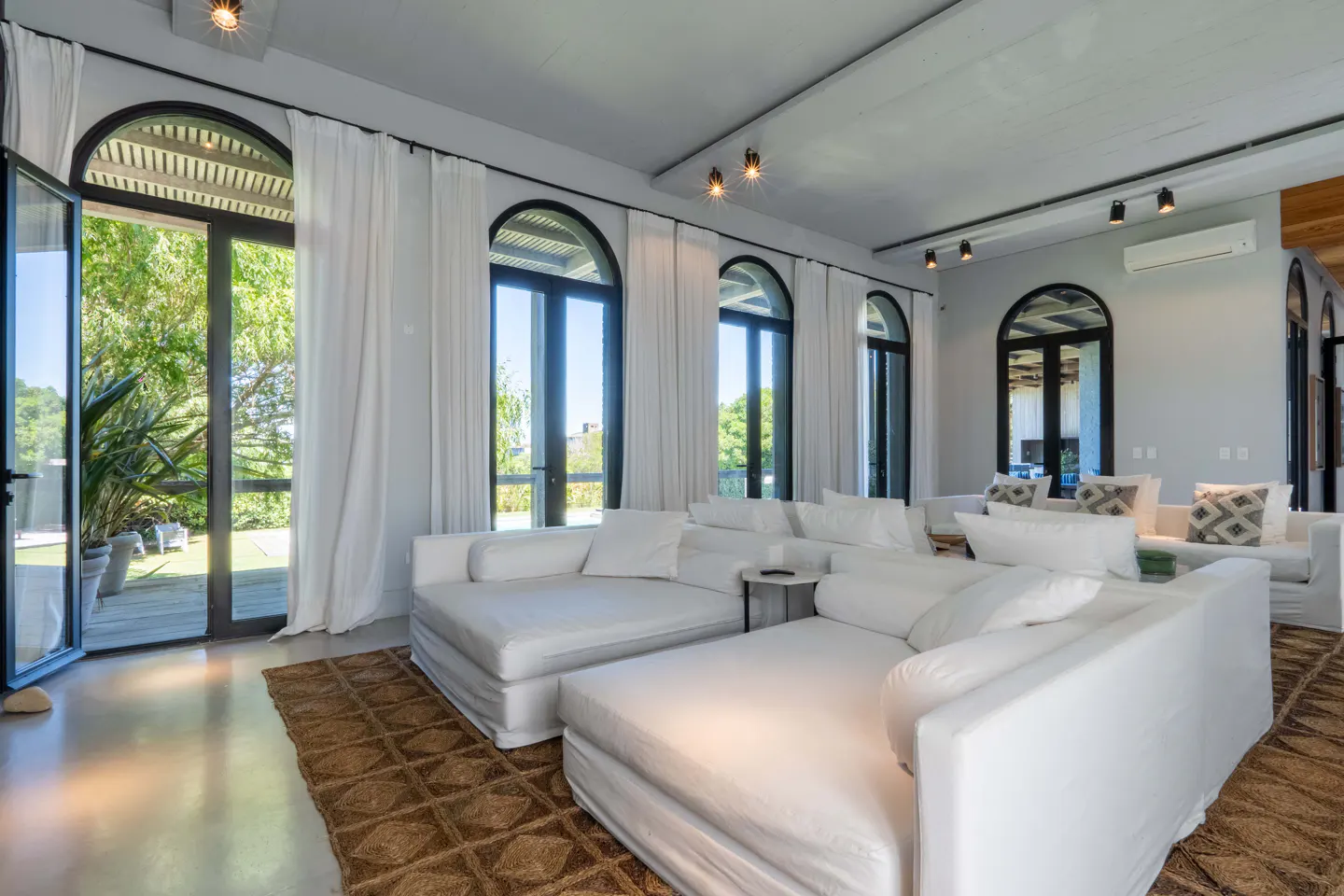 Bright living room with white sofas, arched windows with white curtains, and a brown rug. Outside, green trees and plants are visible.