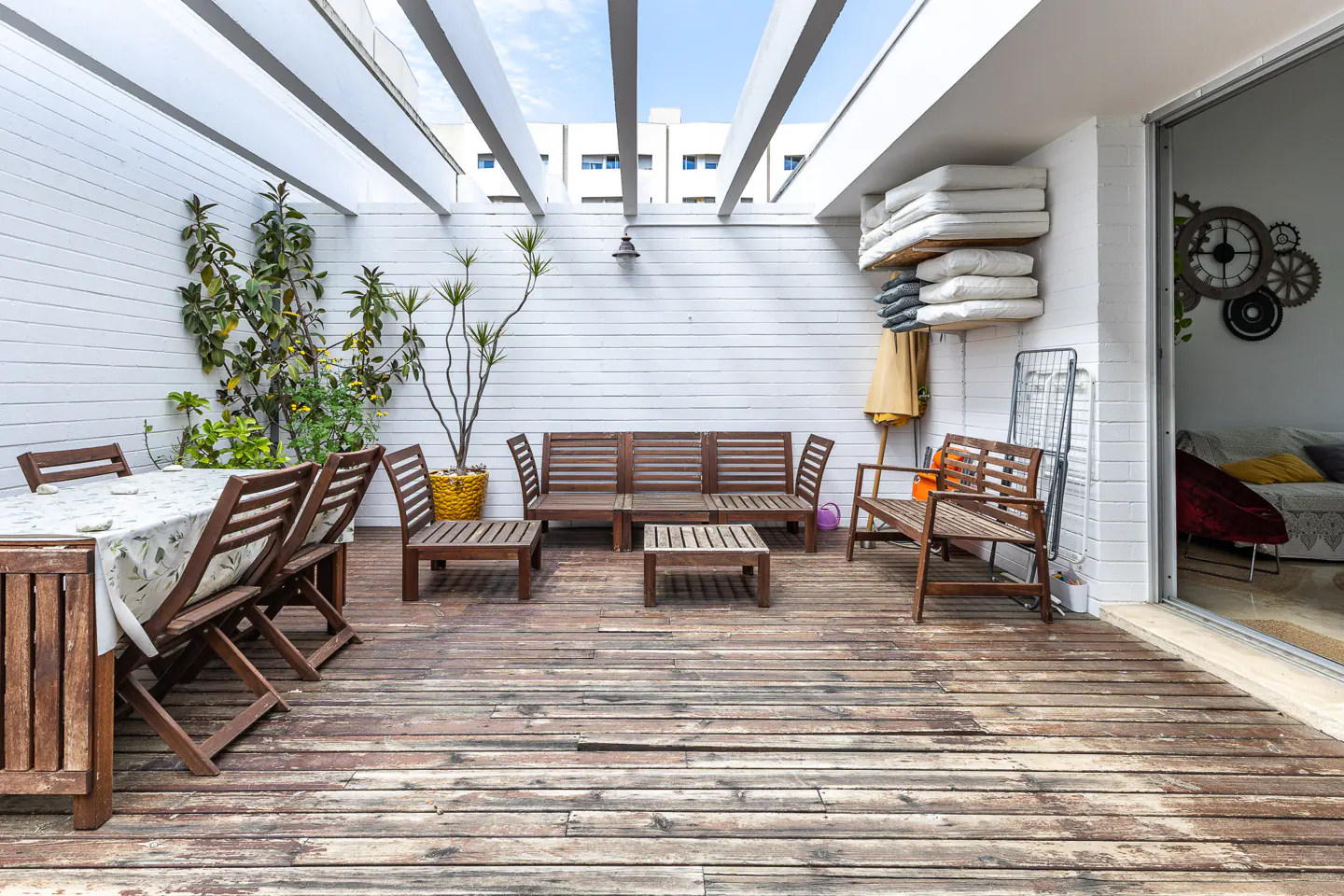Outdoor patio with wooden furniture, plants, and a white pergola. The floor is made of weathered wood planks.