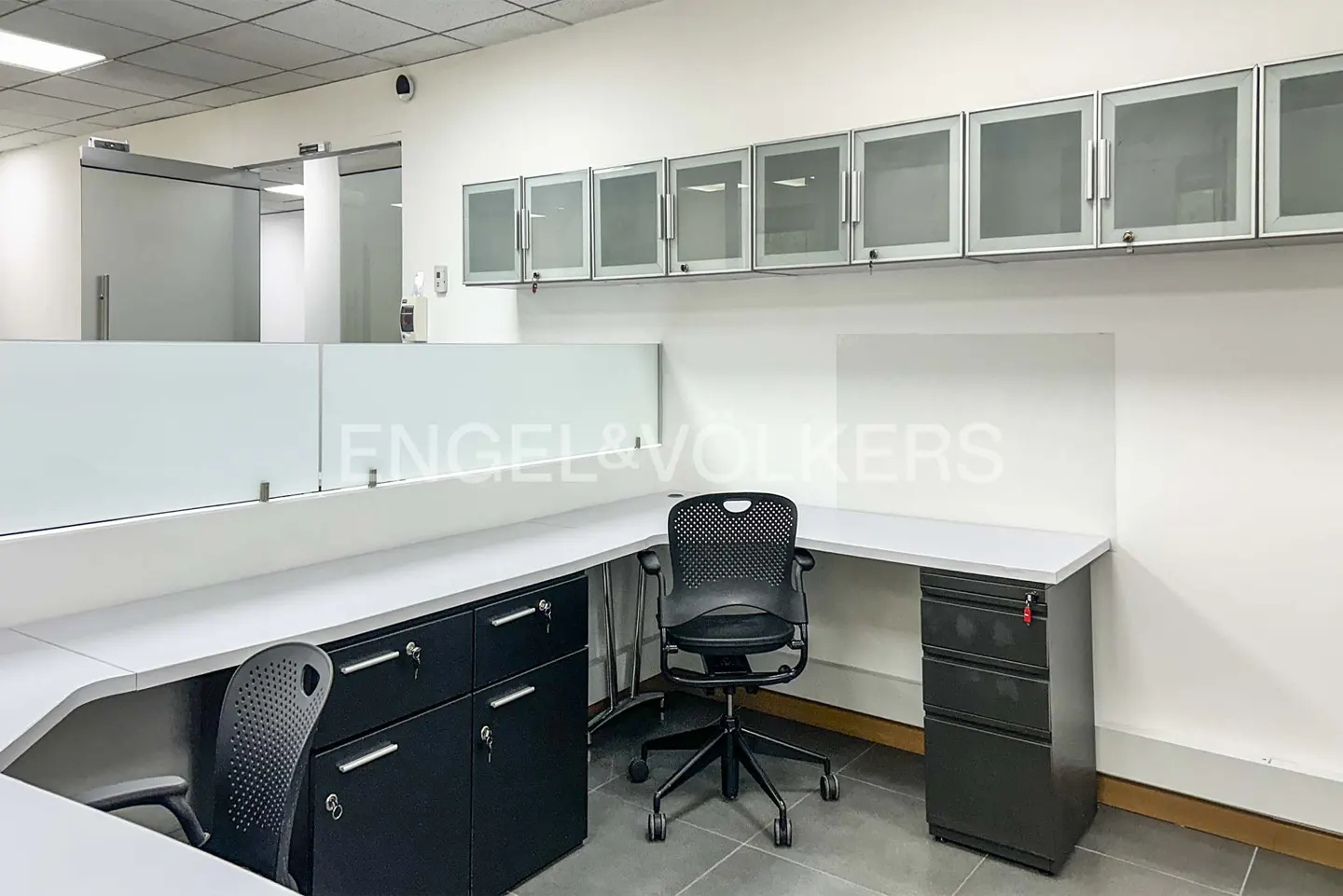 Office space with white walls, gray tile floor, desks, black chairs, and cabinets. Engel & Volkers logo on a glass partition.
