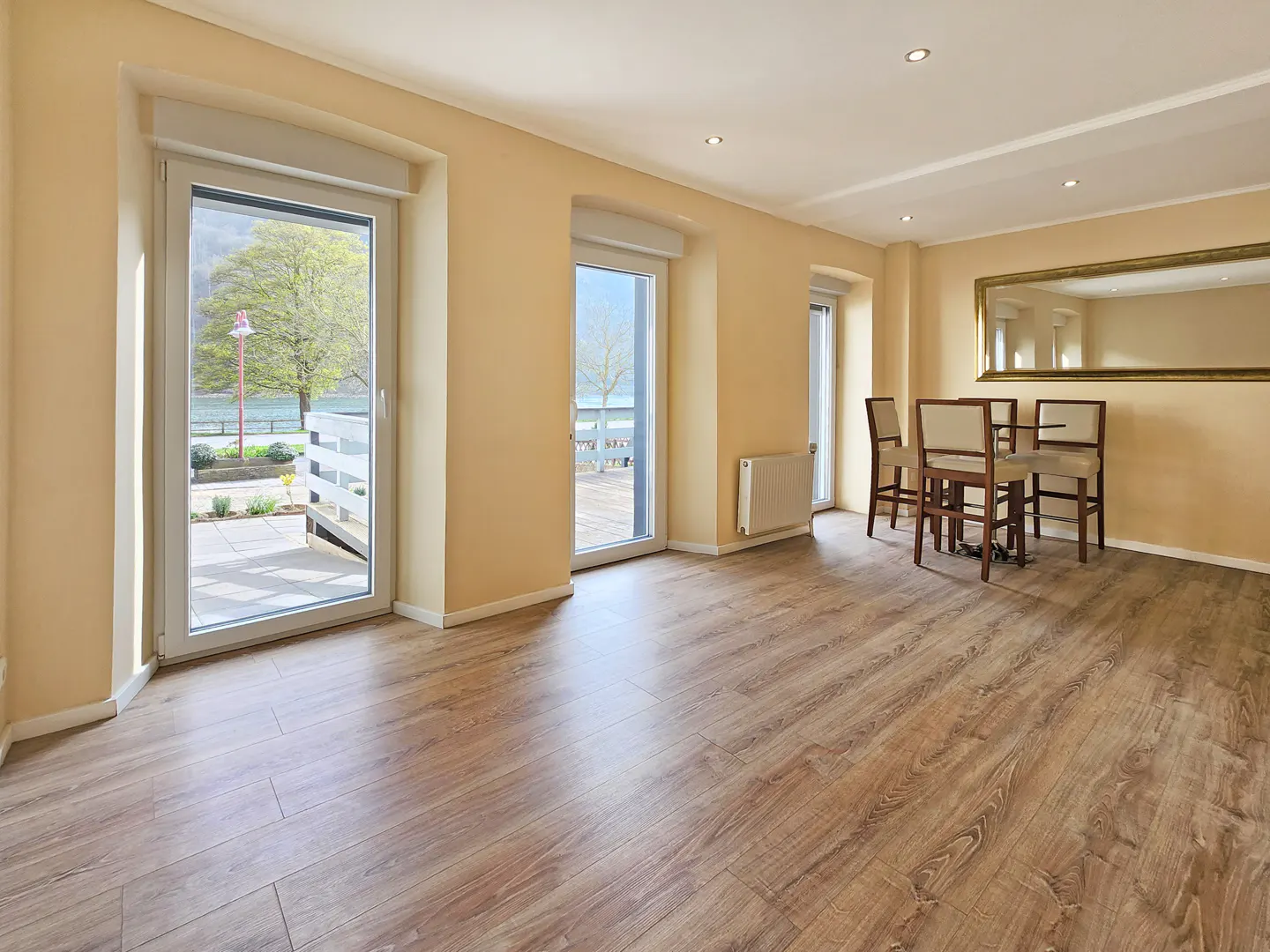Bright, empty dining room with wood floors, beige walls, and a large mirror. Patio doors open to an outdoor space. Table and chairs are set.