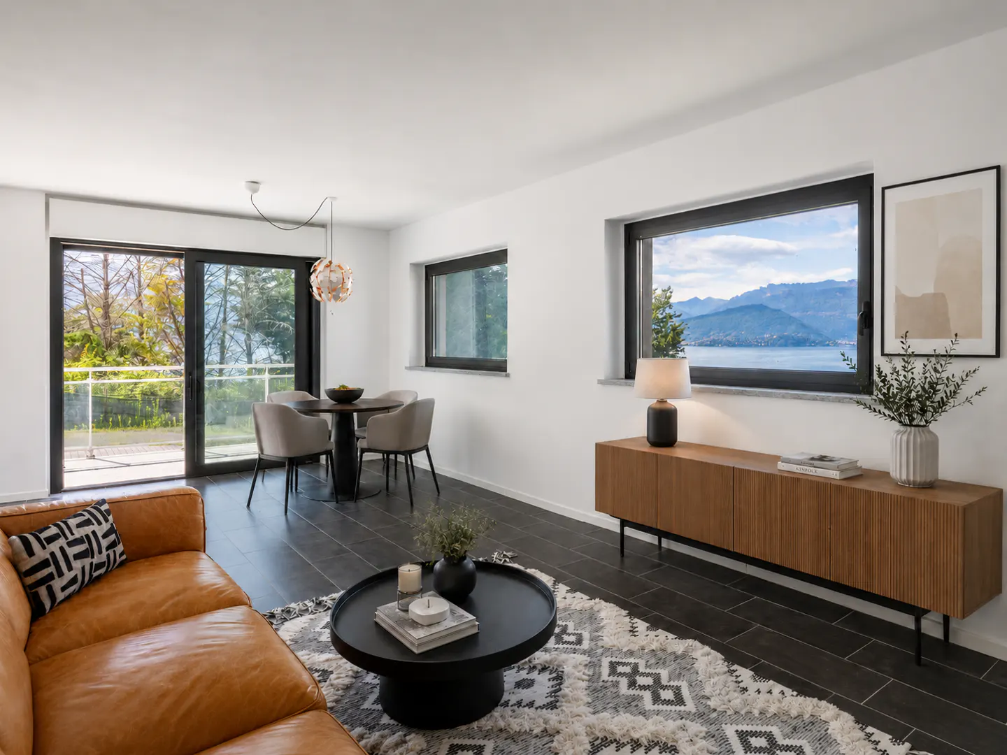 Bright living room with a brown leather sofa, black coffee table, and dining set. A large window shows a lake and mountains.