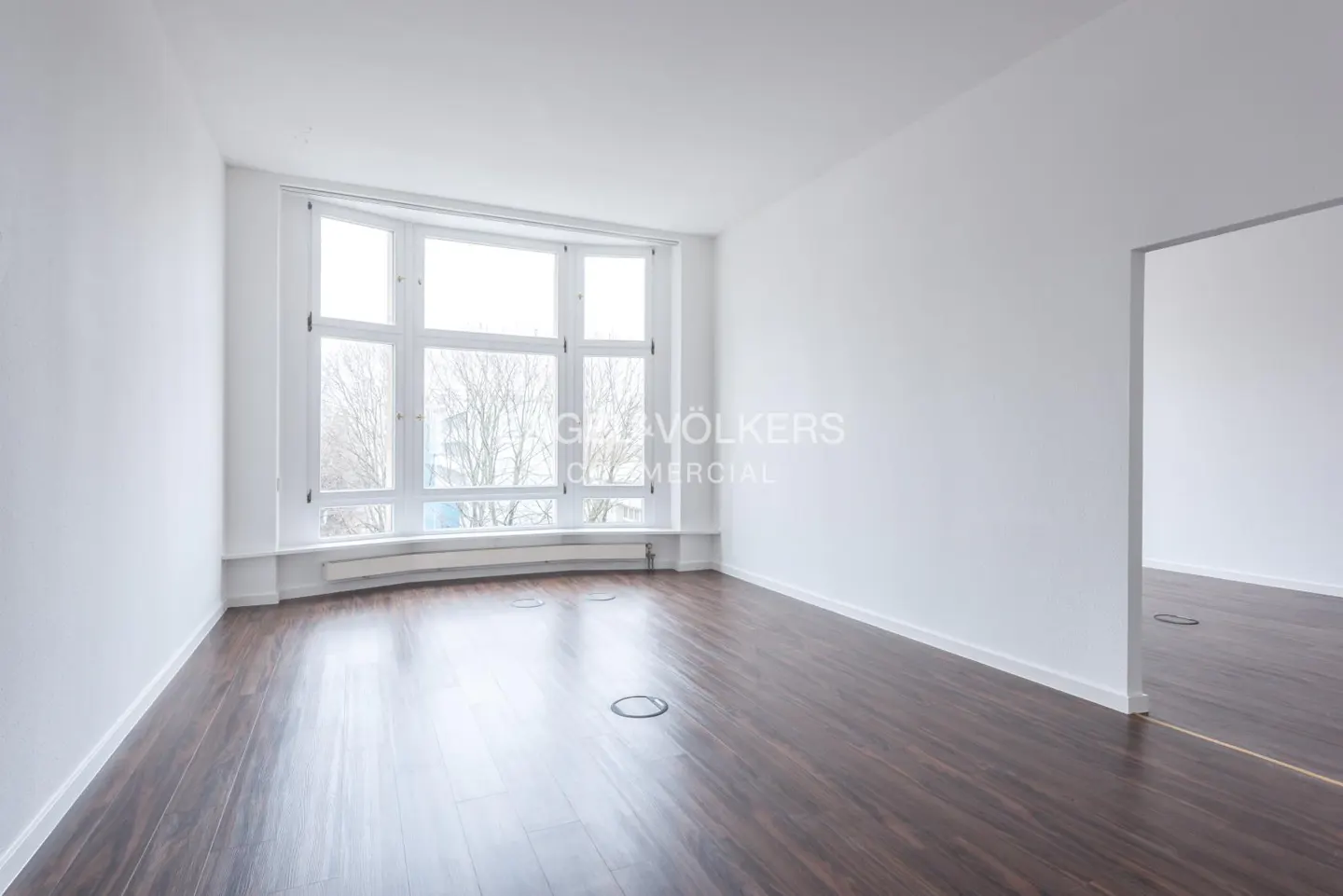Empty room with dark wood floors, white walls, and a large window. Another room is visible through a doorway.