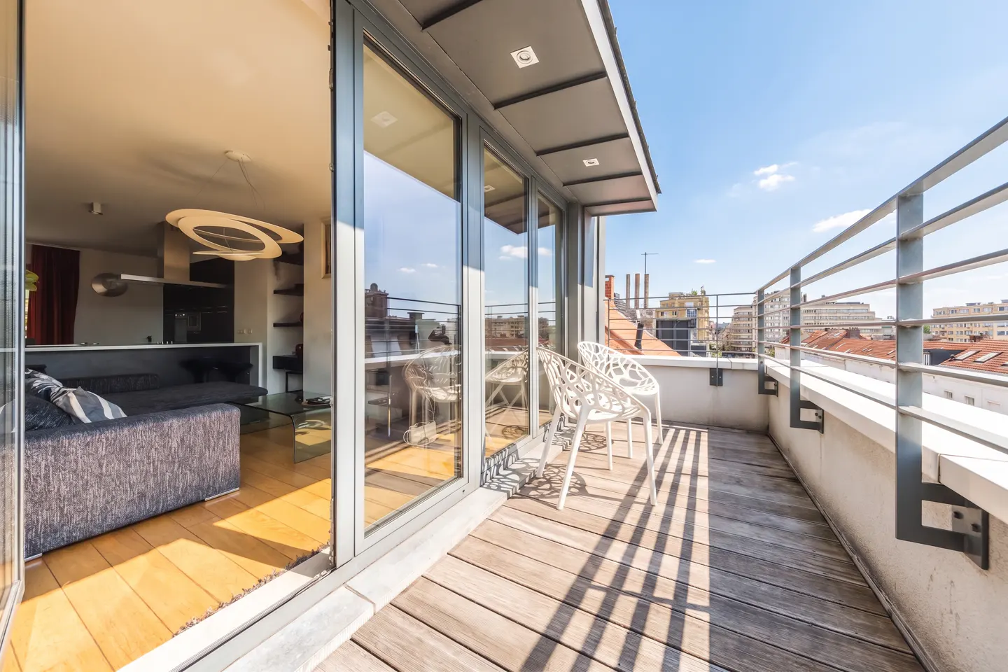 Apartment balcony with wood floor, metal railing, and white chairs overlooking city buildings under a blue sky. Living room visible through glass doors.