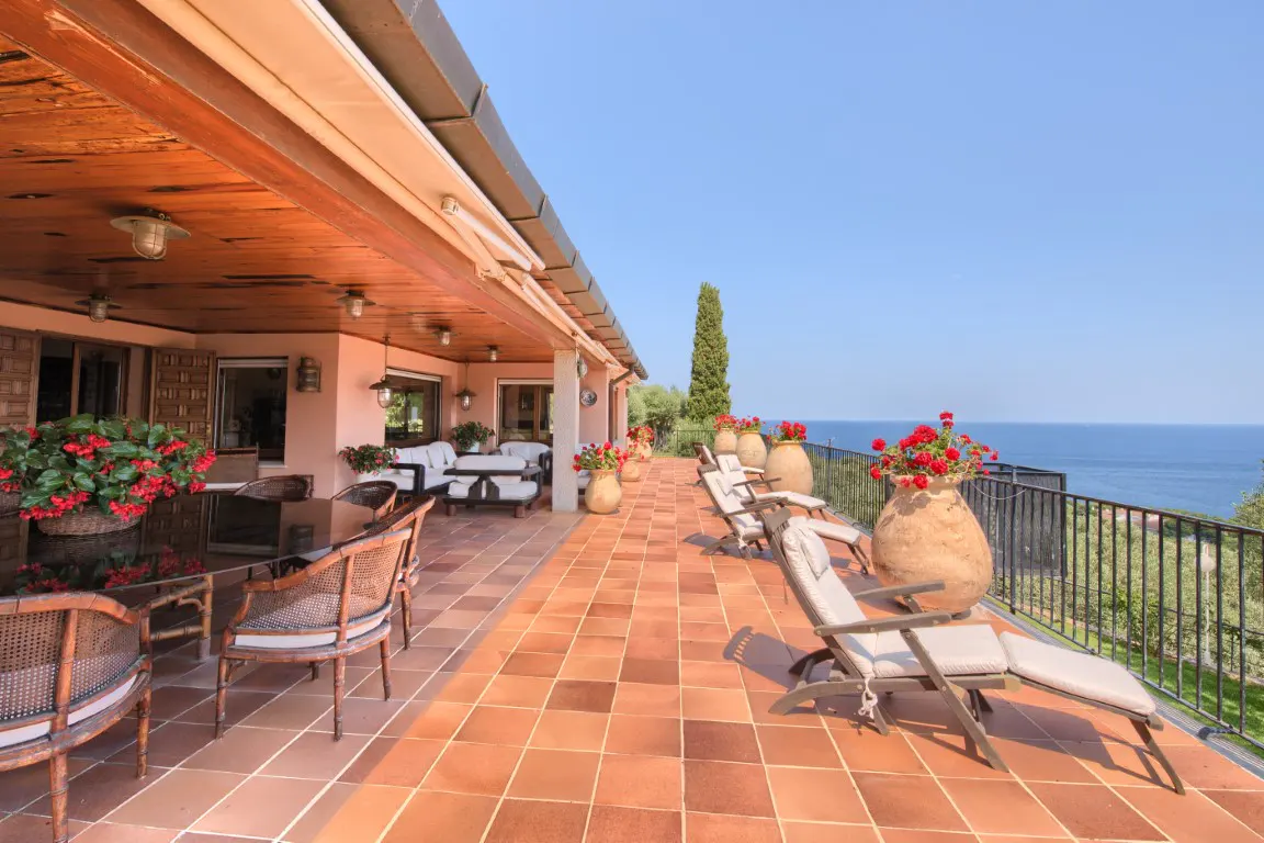 Outdoor patio with terracotta tiles, lounge chairs, and red flowers in pots overlooking the ocean.