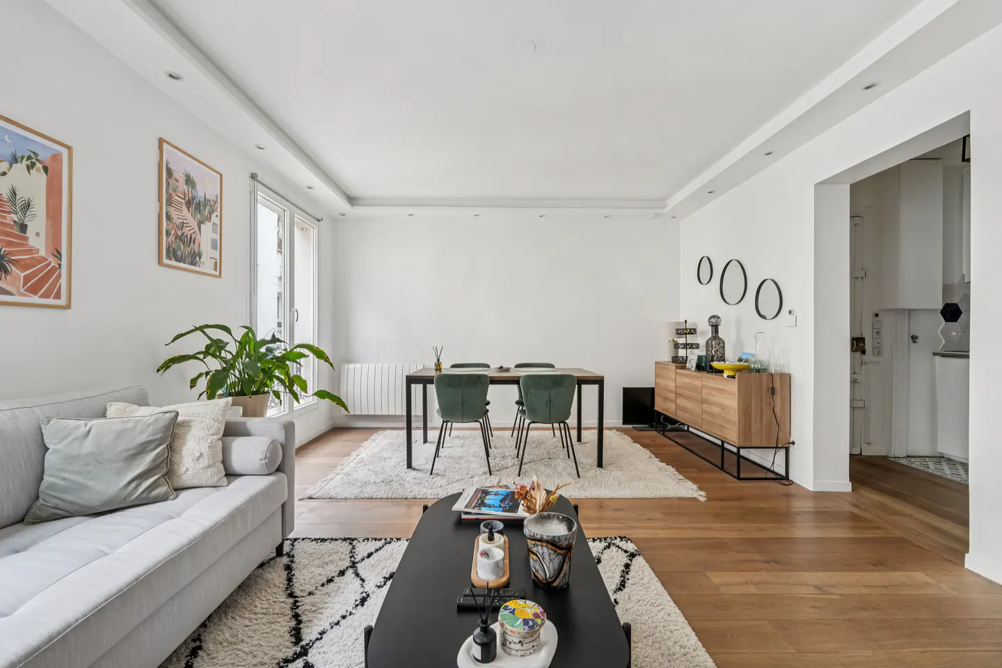 Bright living room with white walls, wood floors, and a gray sofa. A black coffee table sits on a patterned rug. A dining table with green chairs is in the background.
