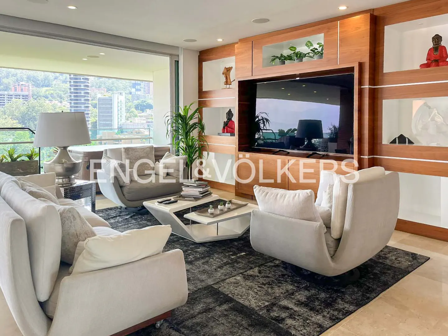 Living room with white sofa, chairs, and a modern coffee table on a dark rug. A large TV is mounted on a wooden wall unit. Balcony with city view.