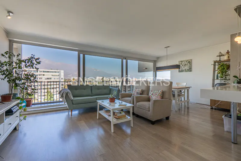 Living room with a green sofa, two beige armchairs, and a white coffee table on a light wood floor. Large windows overlook a city and mountains.