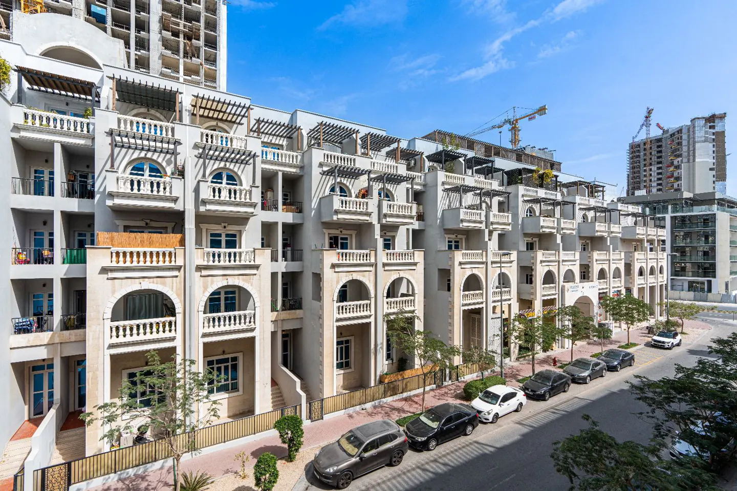 Exterior view of a light gray apartment building with arched balconies and parked cars on the street.