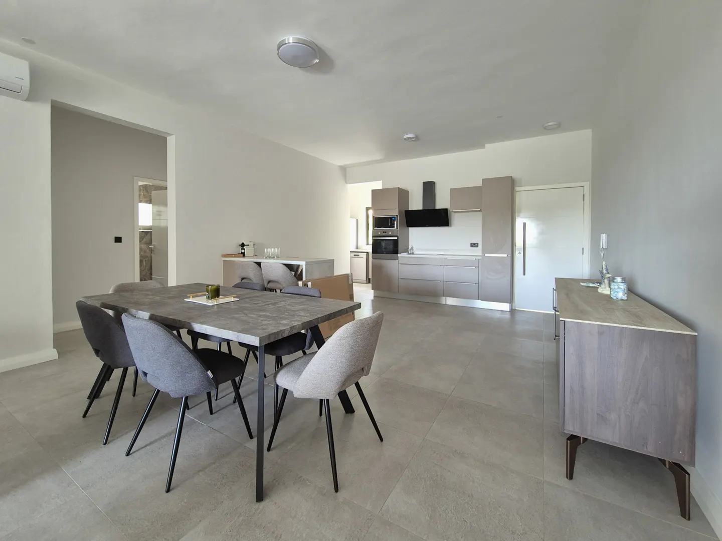 A modern, open-concept dining and kitchen area with gray tile flooring. A dining table with six chairs sits near a kitchen with taupe cabinets.