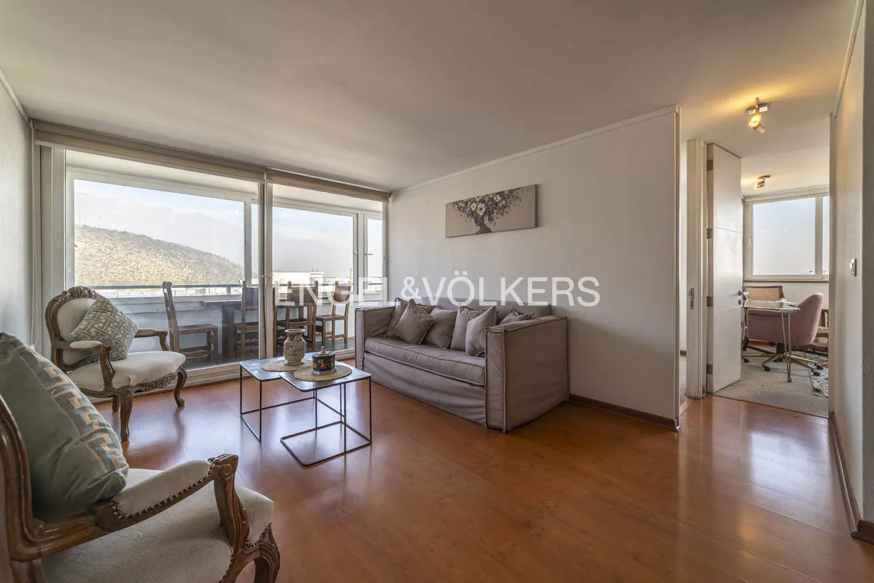 Living room with hardwood floors, beige sofa, two armchairs, and a balcony with mountain views. Engel & Völkers logo on the wall.