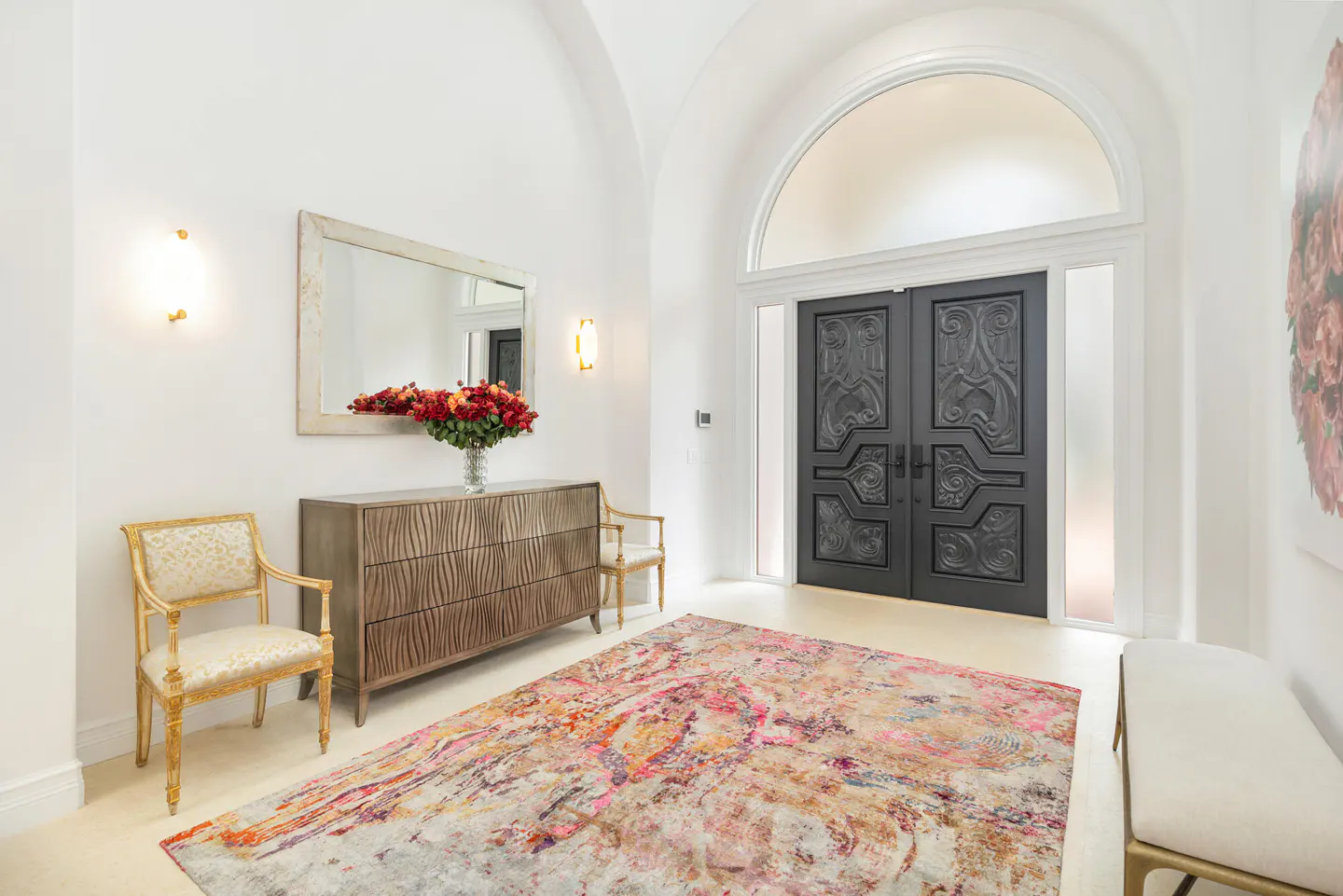 Bright foyer with white walls, arched doorway, and carved black double doors. A colorful rug sits in front of a wooden dresser with flowers and a mirror.