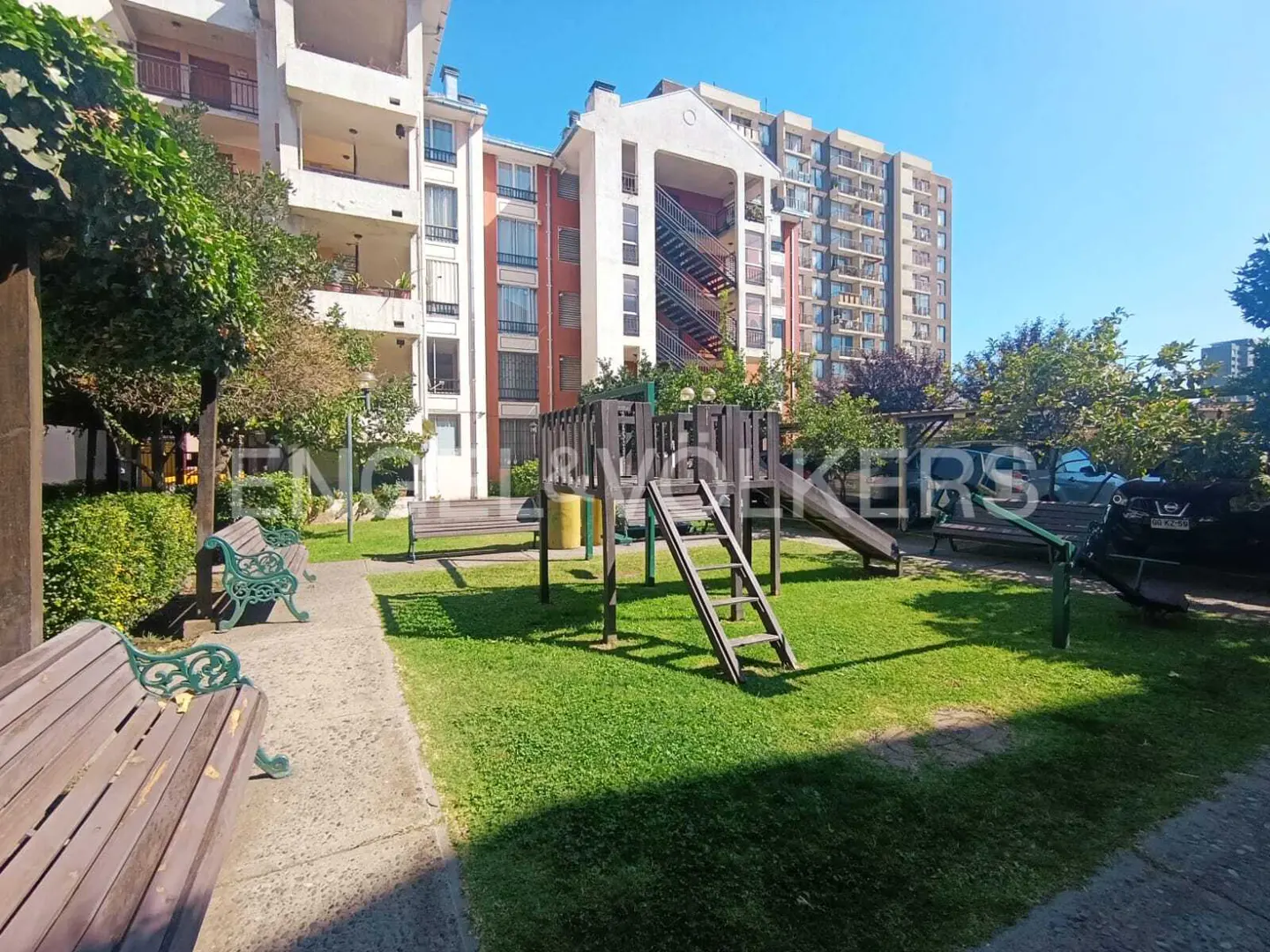Apartment complex with a playground. Green grass, benches, and trees surround the play area. Blue sky above.