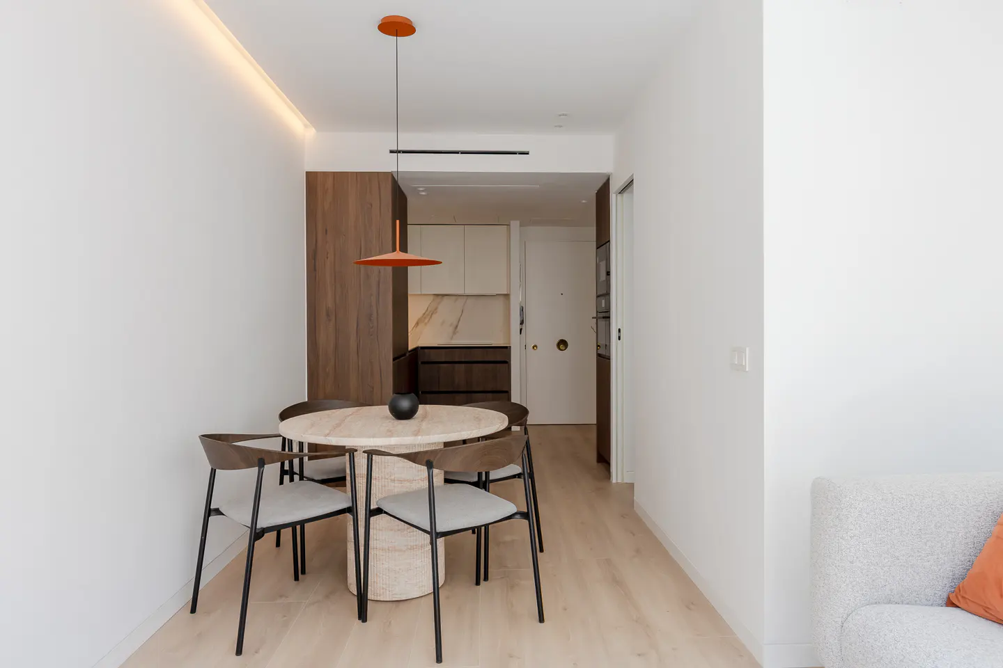 A modern dining area with a round table, four chairs, and an orange pendant light. The kitchen is visible in the background.