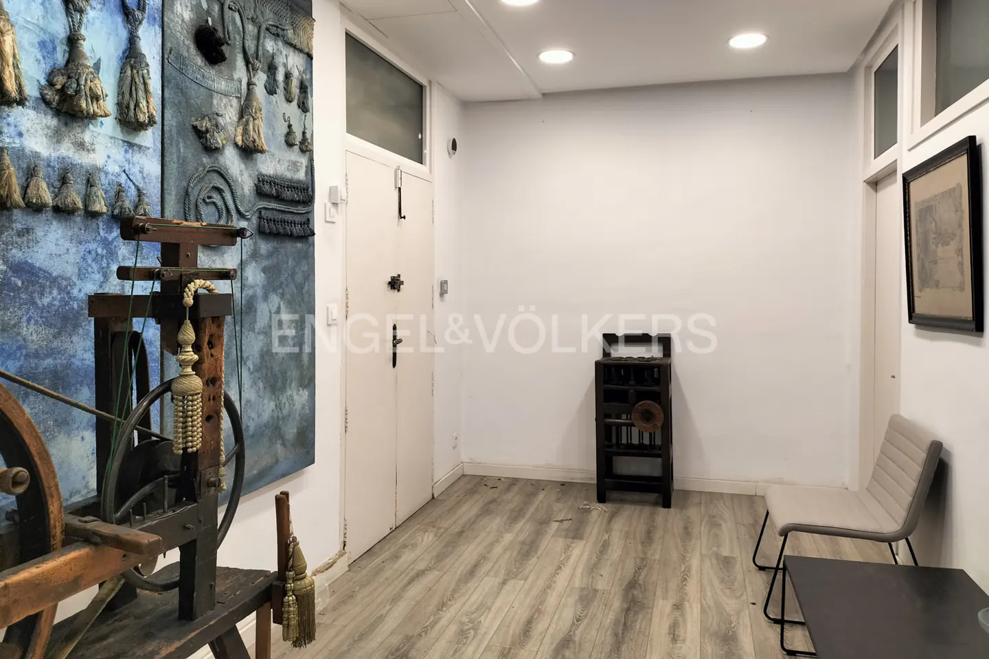 A real estate interior shows a hallway with art, a chair, and a cabinet. The walls are white, and the floor is light wood.