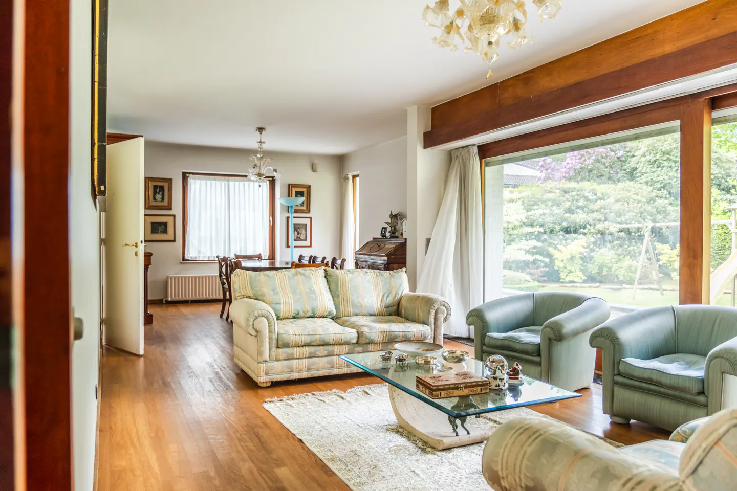 Living room with hardwood floors, patterned sofa, glass table, and green armchairs. Large windows overlook a green yard.