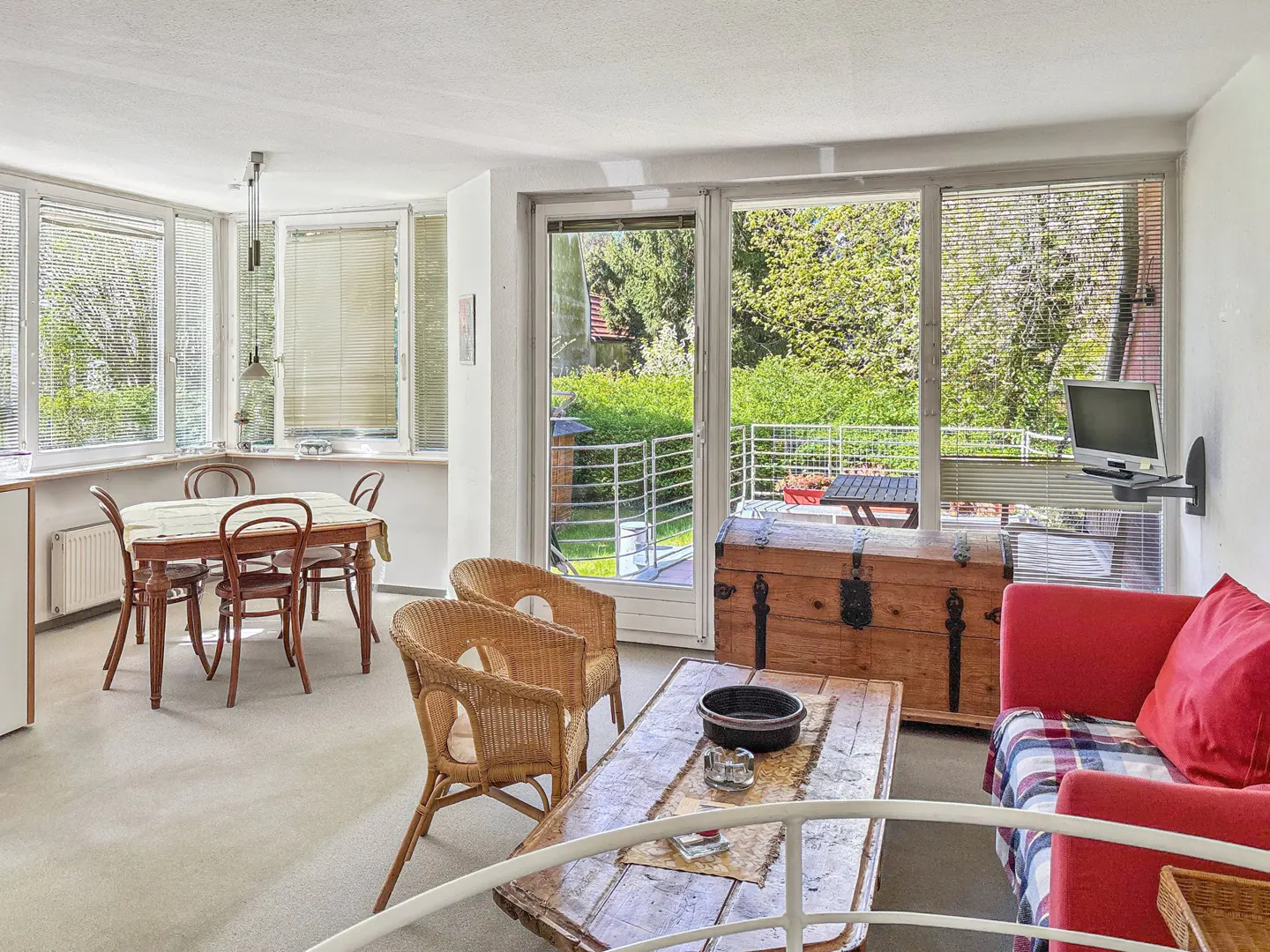Bright living room with dining table, wicker chairs, wooden chest, and red sofa. Balcony view of green trees.
