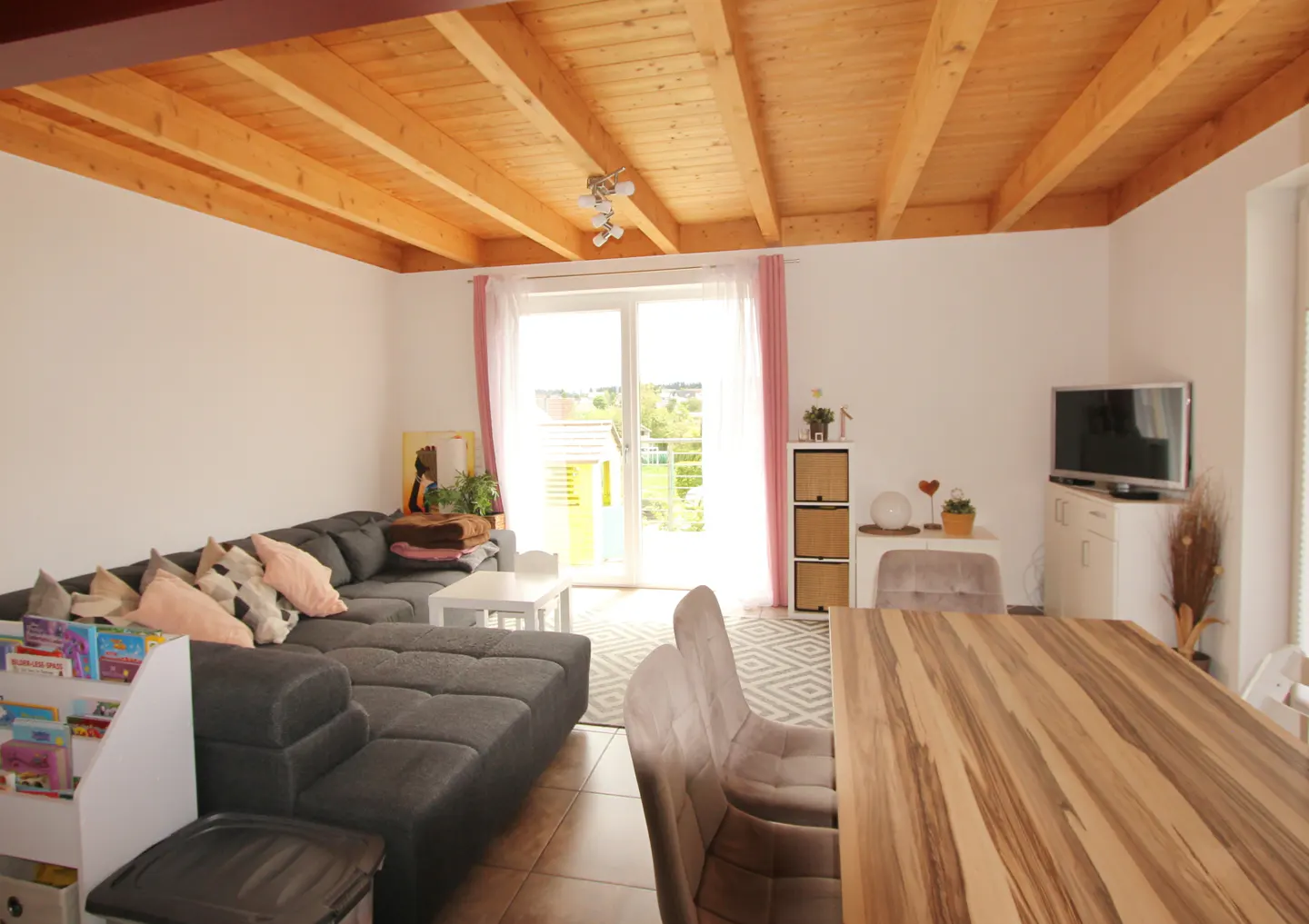 Bright living room with wood ceiling, gray sofa, and dining table. Large windows with pink curtains let in natural light.
