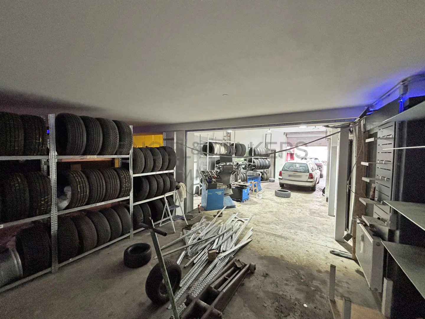 Inside a tire shop, tires are stacked on shelves. A car is visible in the background. Tools and equipment are scattered around.