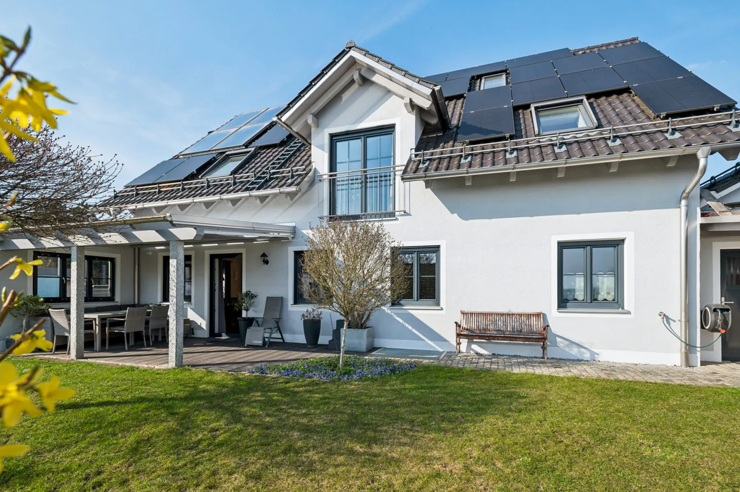 Modern white house with solar panels on the roof, a patio, and a green lawn on a sunny day.