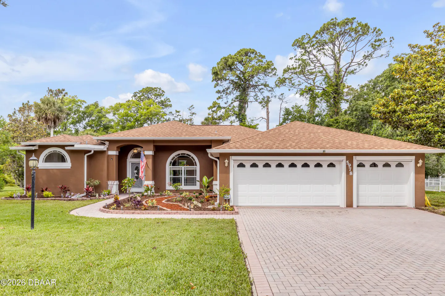Single-story brown stucco home with a tan shingle roof, three-car garage, and landscaped front yard under a blue sky.