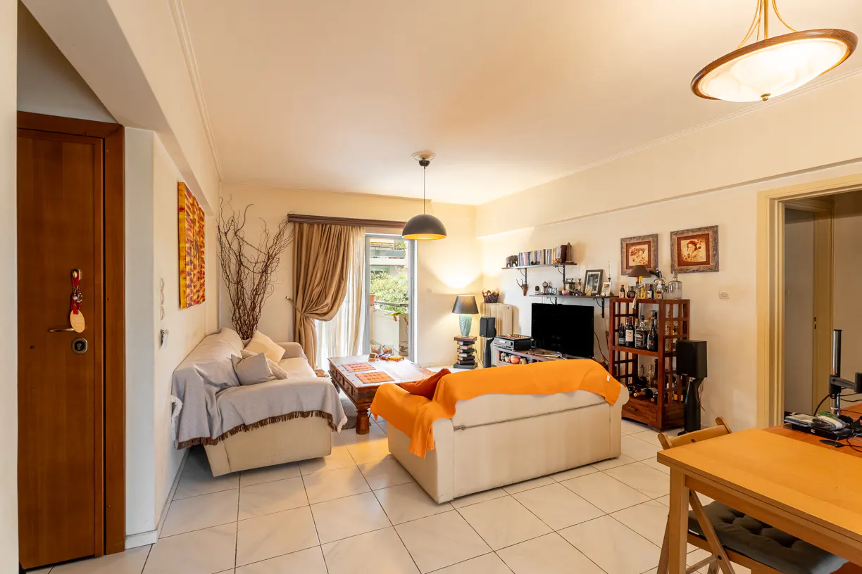 Living room with beige sofas, wood table, TV, and balcony. Neutral colors and tile flooring.