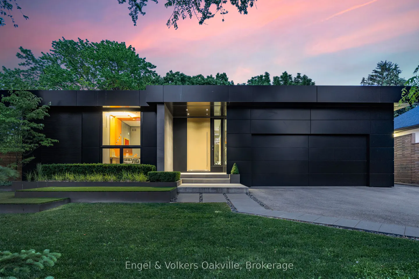 Modern, black, single-story home with a flat roof, green lawn, and pink sky at dusk.