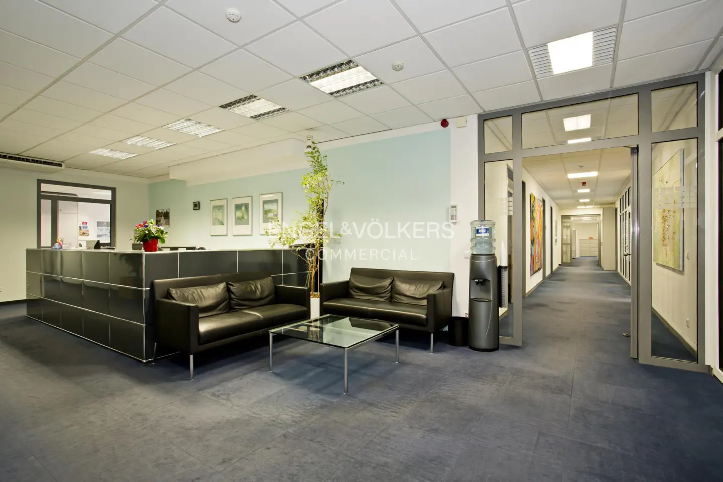 Office lobby with black leather sofas, glass table, and reception desk. A water cooler stands near a hallway entrance.