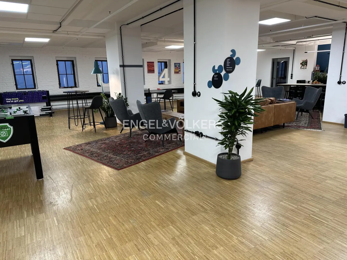 Bright office space with wood floors, lounge area, foosball table, and Engel & Völkers Commercial logo.