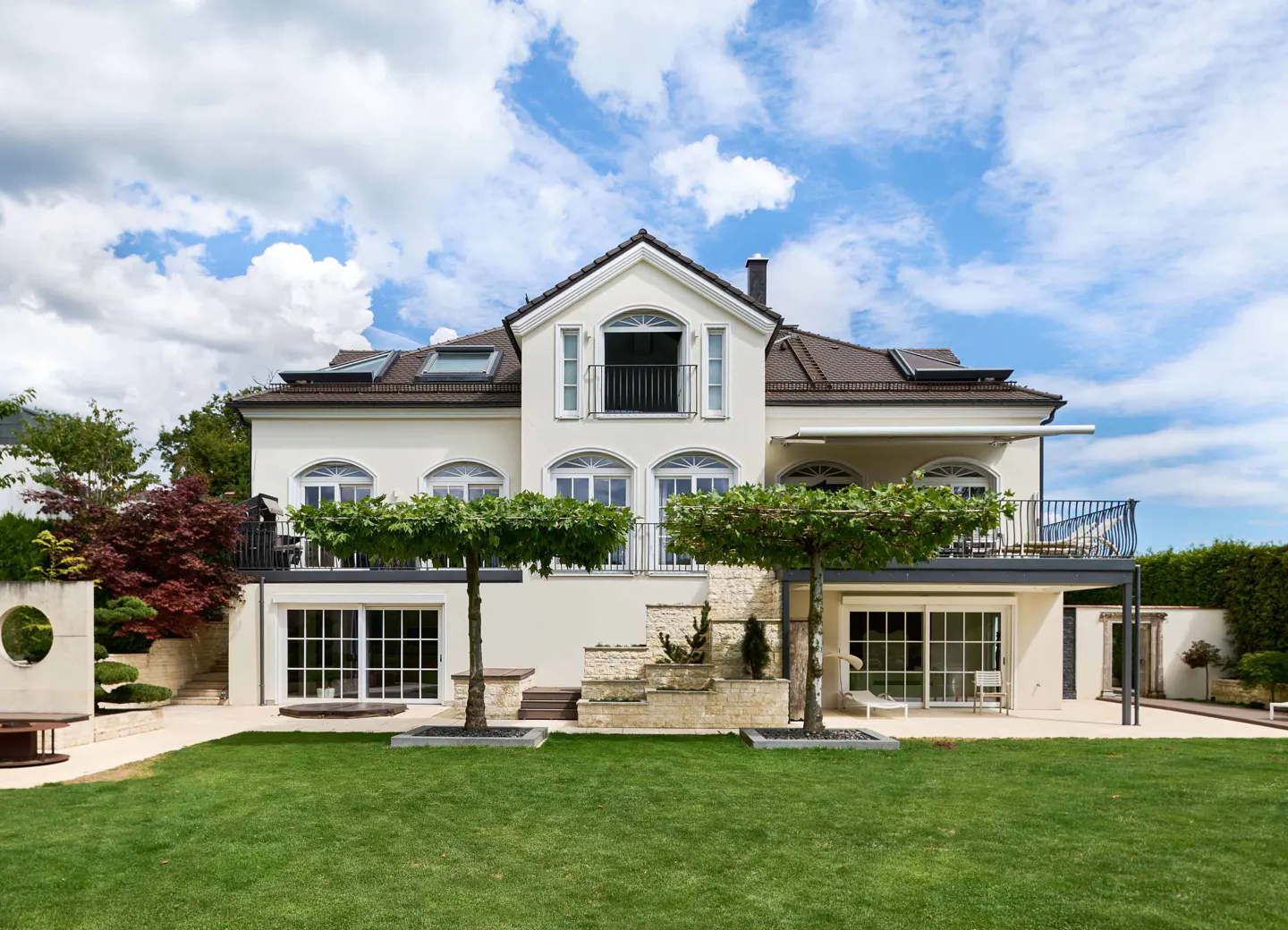 Back view of a large, cream-colored house with a brown roof and green lawn under a blue, cloudy sky.