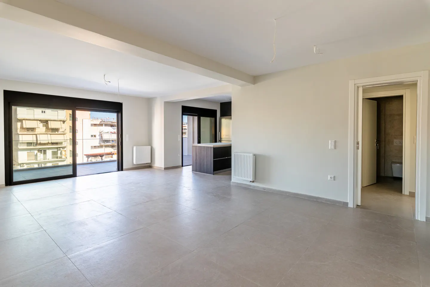 Bright, empty apartment with gray tile floors, white walls, and black-framed sliding doors to a balcony. A kitchen island and bathroom are visible.