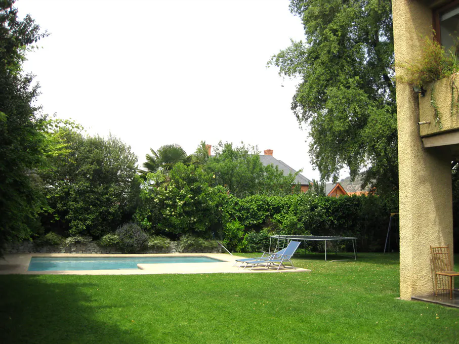 Backyard with a pool, green lawn, trees, and blue lounge chairs. A trampoline is in the background.