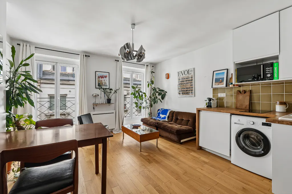 Bright studio apartment with hardwood floors, a brown sofa, dining table, and a kitchen area with a built-in washer.