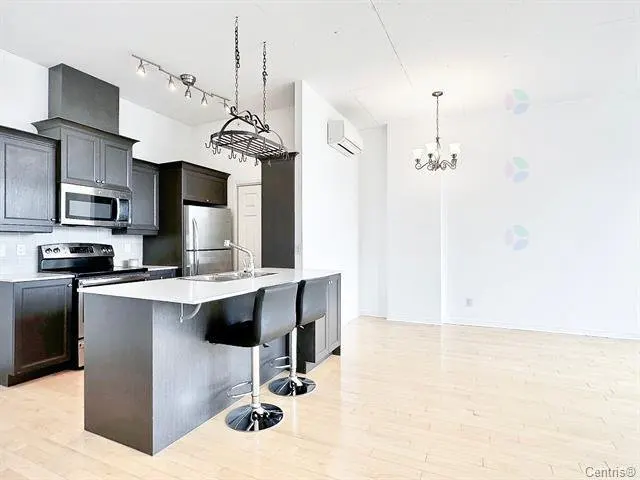 Bright, modern kitchen with gray cabinets, stainless steel appliances, and a white countertop island with two black bar stools. A chandelier hangs over the light wood floor.