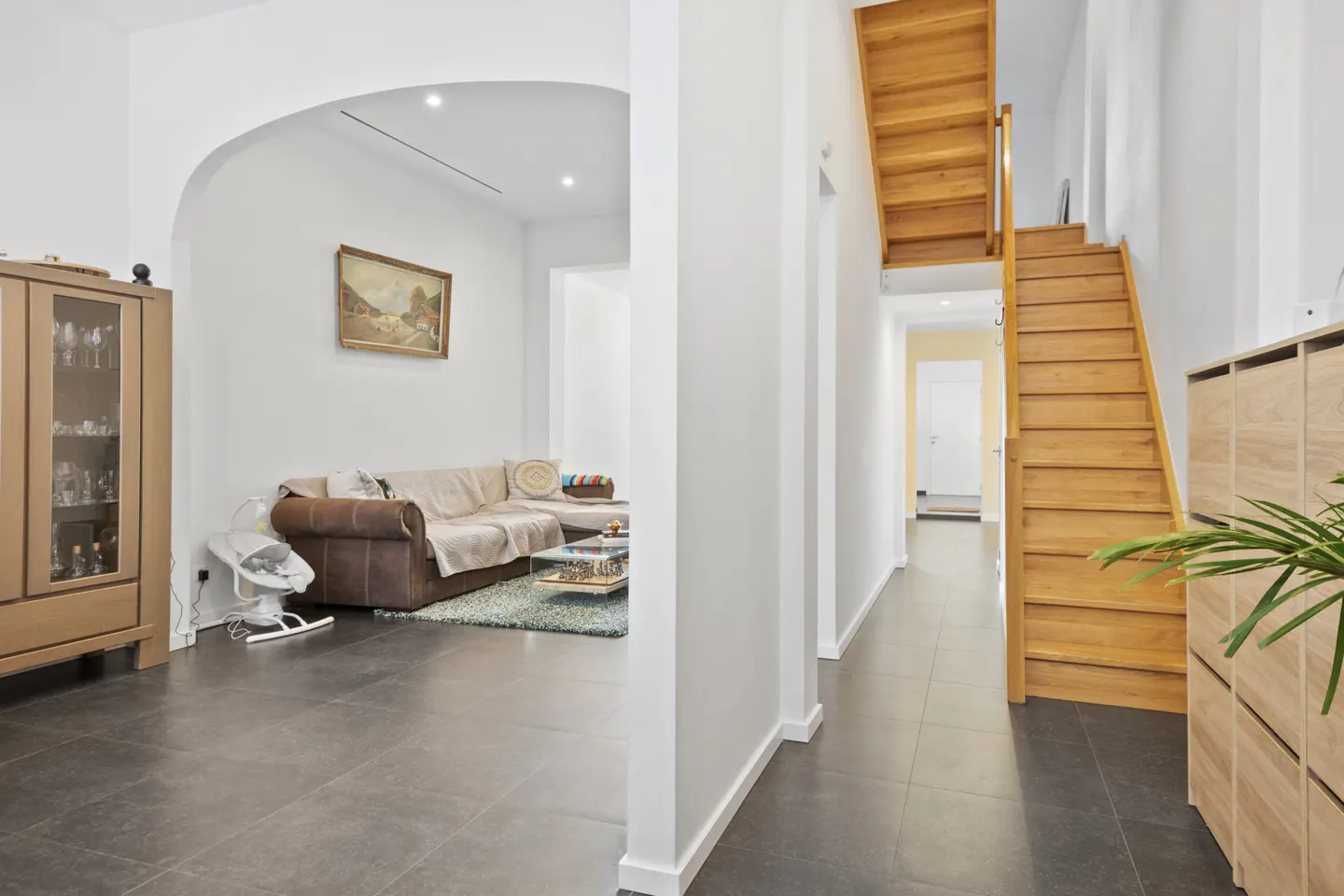 A bright hallway with dark tile floors leads to a wooden staircase, with an arched opening to a living room on the left.