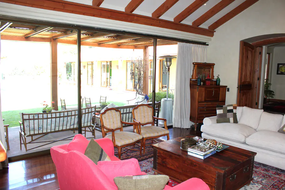A bright living room with a pink sofa, wooden coffee table, and chairs. A large window overlooks a patio with seating and a green lawn.